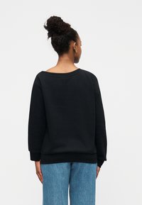 Μαύρο φούτερ με φαρδιά λαιμόκοψη, μακριά μανίκια και ριπ μανσέτες. Συνδυασμένο με ανοιχτόχρωμο μπλε τζιν, η υφή φαίνεται απαλή και ελαφρώς ανάγλυφη.