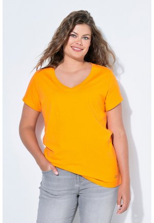 Frau mit locker fallendem, welligem Haar, die ein leuchtend orangefarbenes V-Ausschnitt-T-Shirt und hellgraue Jeans trägt, lächelt und mit einer Hand in der Tasche steht.