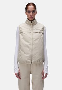 Gilet rembourré beige avec un col haut, fermeture éclair devant et deux poches poitrine horizontales. Superposé sur un haut à manches longues blanc. Texture lisse.