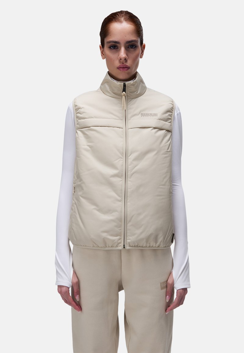 Gilet rembourré beige avec un col haut, fermeture éclair devant et deux poches poitrine horizontales. Superposé sur un haut à manches longues blanc. Texture lisse.