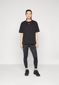 Svart oversized t-skjorte med en liten logo på brystet, kombinert med svarte leggings med teksturerte striper, og svarte joggesko.
