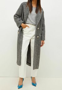 Manteau oversize gris à motif chevrons avec deux boutons, assorti à un pantalon blanc texturé et des escarpins bleus à bouts pointus.