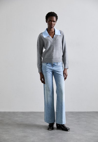 Suéter gris de cuello en V con un color azul claro, combinado con jeans de pierna ancha azul claro y botas de tacón negro, de pie sobre un suelo gris.