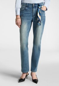 Jeans in denim azzurro chiaro con vestibilità comoda, leggermente sbiaditi, tasche anteriori e una sciarpa di seta legata in vita. Scarpe nere con tacco alto.