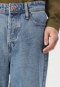 Hellblaue Jeans aus Denim mit regulärer Passform, ausgestattet mit einem Kupferknopf, sanften Ausbleichungen und einem Fünf-Taschen-Design. Hand ruht auf der Seitennaht.