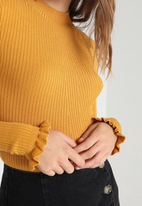 Sinine rib wzorze musztardowym z wysokim dekoltem i falbankami przy mankietach. Połączony z czarną spódnicą z ozdobnymi guzikami. Widoczne długie rękawy.