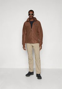 Rudas flisinis švarkelis su užtrauktuku, juodais akcentais ir logotipu. Derinamas su smėlio spalvos kelnėmis ir juodais batais. Modelis dėvi saulės akinius.