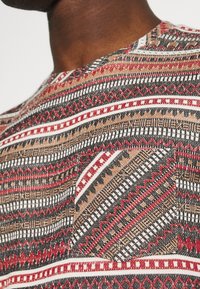Camicia a maniche corte con un motivo tribale multicolore in rosso, nero, bianco e marrone, con un dettaglio di tasca sul lato sinistro.