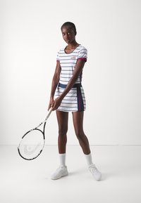 Vestido de tenis blanco a rayas con acentos en azul marino y rojo, mangas cortas y cintura ajustada; la modelo sostiene una raqueta de tenis.