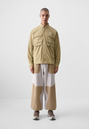 Khaki koszula zapinana na guziki z czterema przednimi kieszeniami, w połączeniu z szerokimi spodniami w kolorach beżowym i szarym. Model nosi szare sneakersy.