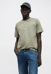 T-shirt verde oliva a maniche corte con logo frontale, abbinata a jeans di denim blu; modella con le mani nelle tasche, indossa un berretto scuro.