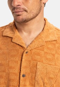 Textured orange collared shirt featuring a checkered pattern with circular accents, button closure, and a chest pocket.