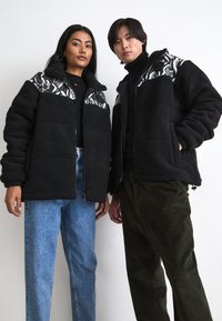 Fuzzy black jackets featuring patterned shoulders, zip closure, and pockets. Models wear light blue jeans and dark green corduroy trousers.
