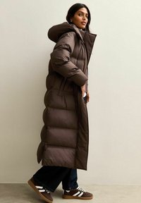 Abrigo tipo puff marrón con capucha, que presenta un diseño largo, acolchado y con patrón de rombos. Combinado con jeans oscuros y zapatillas marrones.
