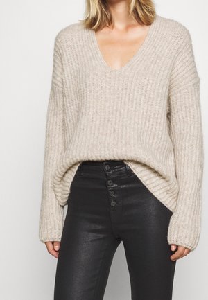 Kvinde iført en beige ribbet V-hals sweater stoppet ned i højtaljede sorte bukser med knapper foran, står mod en ensfarvet baggrund.