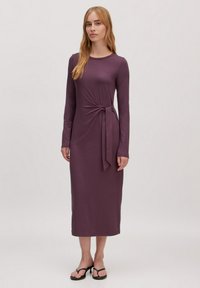 Robe longue mauve à manches longues avec un détail de taille nouée, faite d'un matériau doux et extensible. Longueur au sol avec un col rond.