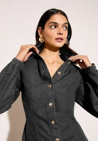 Mustat farkkukangasta oleva mekko, jossa on istuva yläosa, pitkät hihat, napitettu etuosa ja korostuvat kultaiset napit. Sileä tekstuuri ja jämäkät saumat.