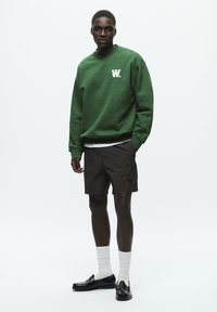 Green sweatshirt with white logo, paired with dark brown shorts. Model wearing white ribbed socks and black loafers. Plain background.