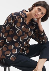 Camicia con bottoni di color blu scuro con grandi motivi geometrici marroni e crema. Tessuto semi-trasparente, maniche lunghe e colletto classico.