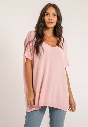 Femme aux longs cheveux noirs portant un haut ample à manches courtes rose clair et un jean bleu, debout devant un fond clair uni.