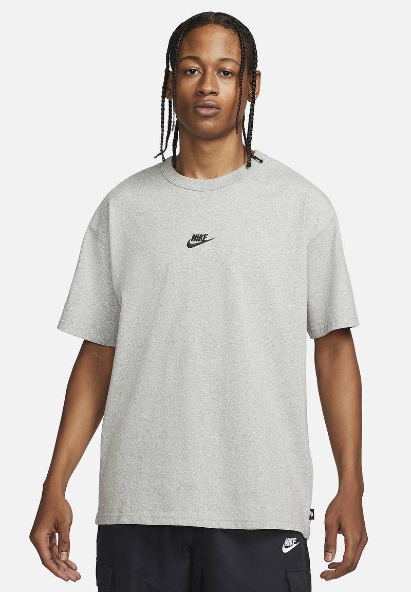 Jaunietis ar pītām matēm, kas valkā gaiši pelēku Nike T-kreklu un melnas Nike šortus uz vienkārša balta fona.