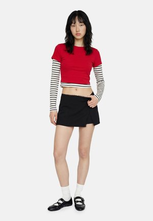 Jonge vrouw draagt een rood cropped topje over een zwart-wit gestreepte longsleeve, een zwarte minirok, witte sokken en zwarte Mary Jane schoenen.