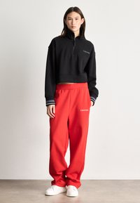 Cropped black sweatshirt with high collar and zipper, paired with loose red sweatpants. Both feature "Calvin Klein" branding. White sneakers.