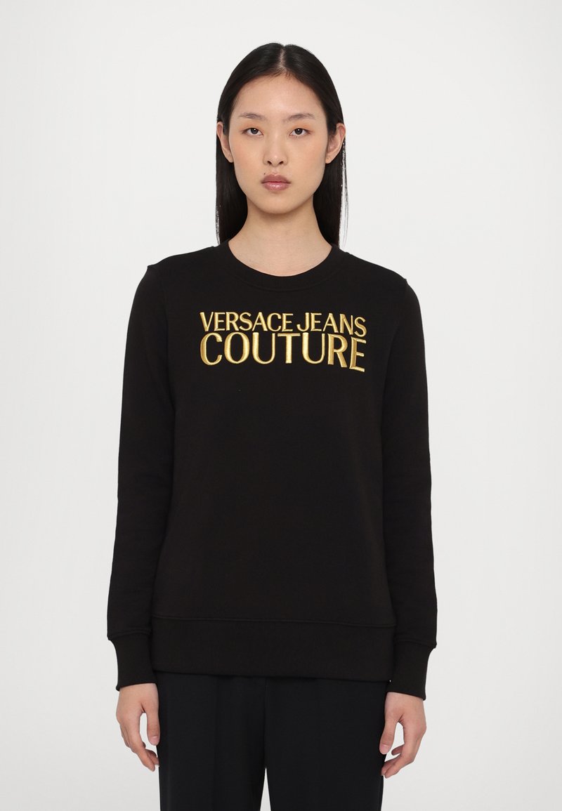 Melna sudraba krekls ar garām piedurknēm, apaļu kakla izgriezumu un zeltīti izšūtu "VERSACE JEANS COUTURE" tekstu priekšpusē. Gluda auduma tekstūra.