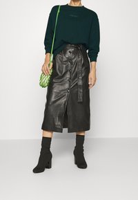 Donna che indossa un maglione verde scuro, una gonna midi in pelle nera con bottoni e cintura, stivali con tacco neri e una borsa a tracolla in rete verde brillante.