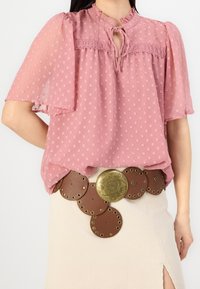 Blouse rose à pois avec des manches bouffantes et un col avec noeud, superposée sur une jupe beige à côtes avec une découpe et une ceinture décorative marron et dorée.