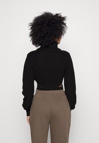 En person med lockigt hår vänder ryggen mot oss, iförd en svart polotröja och beige byxor. Bakgrunden är helt vit.