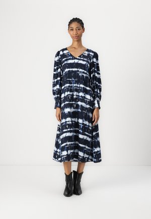 Langes, marineblaues Kleid mit weißem Batikmuster; hat einen V-Ausschnitt, lange Ärmel und eine fließende Silhouette. Kombiniert mit schwarzen Knöchelstiefeln.
