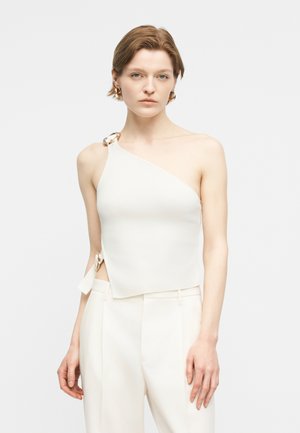 Γυναίκα με κοντά μαλλιά, φοράει λευκό τοπ ενός ώμου με χρυσές λεπτομέρειες σε μορφή κρίκων και λευκό παντελόνι με ψηλή μέση, μπροστά σε απλό φόντο.