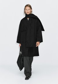 Black overcoat with a wraparound scarf, paired with gray trousers and black shoes, featuring a smooth texture and minimalist design.