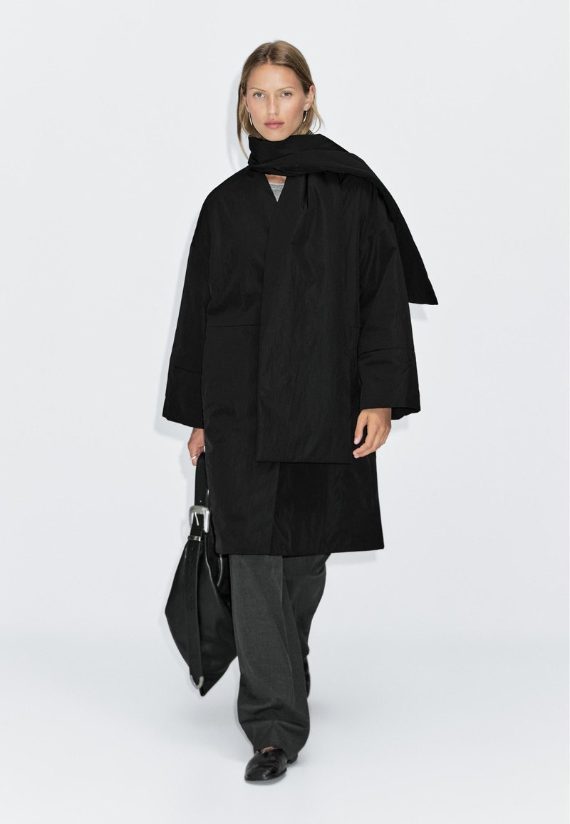 Black overcoat with a wraparound scarf, paired with gray trousers and black shoes, featuring a smooth texture and minimalist design.