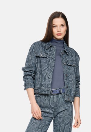 Woman wearing a blue-gray snake print denim jacket and matching pants with a blue belt and dark turtleneck top, standing against a plain background.