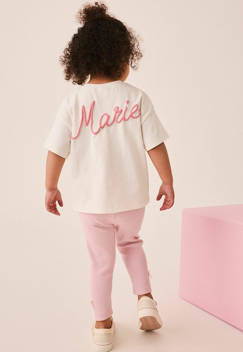 Niño pequeño con cabello rizado caminando, vestido con camiseta blanca con texto rosa "Marie", pantalones rosas y zapatos blancos, cerca de un cubo rosa.