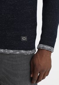 Pull en maille bleu marine avec tissu texturé, bordure rayée blanche et grise, et étiquette noire avec texte blanc en bas.