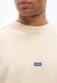 Gros plan sur une personne portant un pull en tricot beige avec un patch logo « HUGO » bleu sur la poitrine.