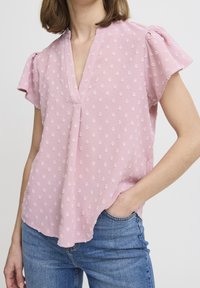 Jasnoróżowa bluzka z krótkimi bufkami, dekoltem w kształcie litery V i fakturą przypominającą kwiaty. Zestawiona z niebieskimi dżinsami.