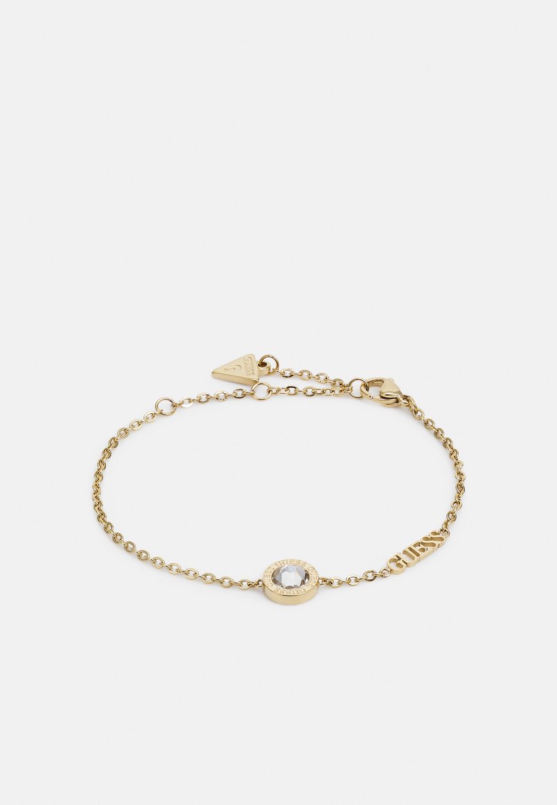 Bracelet en chaîne dorée avec un pendentif circulaire orné d'une pierre précieuse transparente, accentué par le mot "GUESS" en lettres 3D. Fermeture réglable.