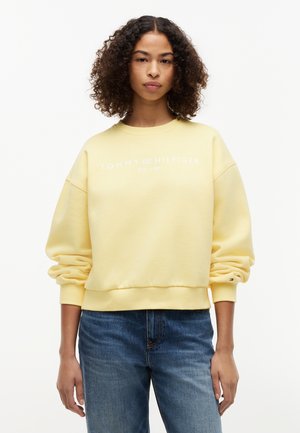 Kvinne med krøllete hår iført en lys gul Tommy Hilfiger-genser og blå jeans, stående foran en hvit bakgrunn.