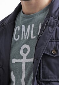 Navy puffer jacket with a zipper and side pocket layered over a green graphic t-shirt featuring a white anchor design.