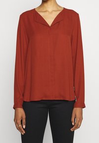 Blouse en tissu léger de couleur rouille à manches longues, col en V, détails de coutures subtiles, coupe décontractée, associée à un pantalon noir.