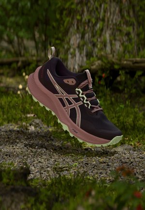 Chaussure de trail noire et rose avec une semelle rugueuse en l’air au-dessus d’un sentier rocheux avec un feuillage vert en arrière-plan.