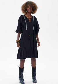 Zwart jurk met een gelaagde rok, pofmouwen en decoratieve afwerking. Heeft een trekkoord met kralen en een kwastje. Draagt met zwarte laarzen.