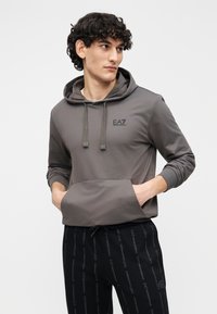 Sivá mikina s kapucňou s predným vreckom a nastaviteľnými šnúrkami, s malým logom EA7. Nosená s čiernymi nohavicami s opakujúcim sa logom značky.