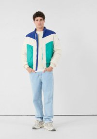 Uomo che indossa un giubbotto imbottito color block bianco, teal e blu, jeans azzurri chiari e sneakers bianche, in piedi contro uno sfondo bianco con le mani in tasca.