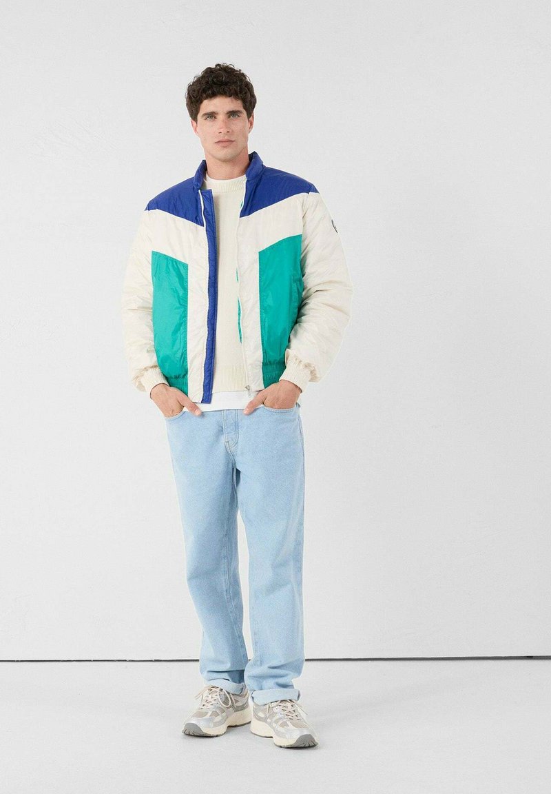 Uomo che indossa un giubbotto imbottito color block bianco, teal e blu, jeans azzurri chiari e sneakers bianche, in piedi contro uno sfondo bianco con le mani in tasca.