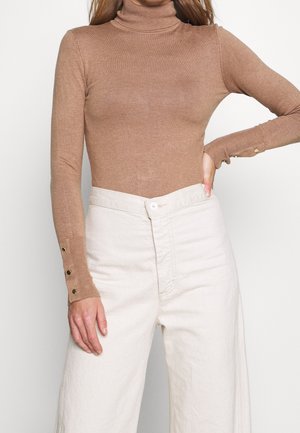 Frau trägt einen eng anliegenden camel-farbenen Rollkragenpullover mit Knopfdetails an den Bündchen und hoch taillierte, off-white weite Hosen.